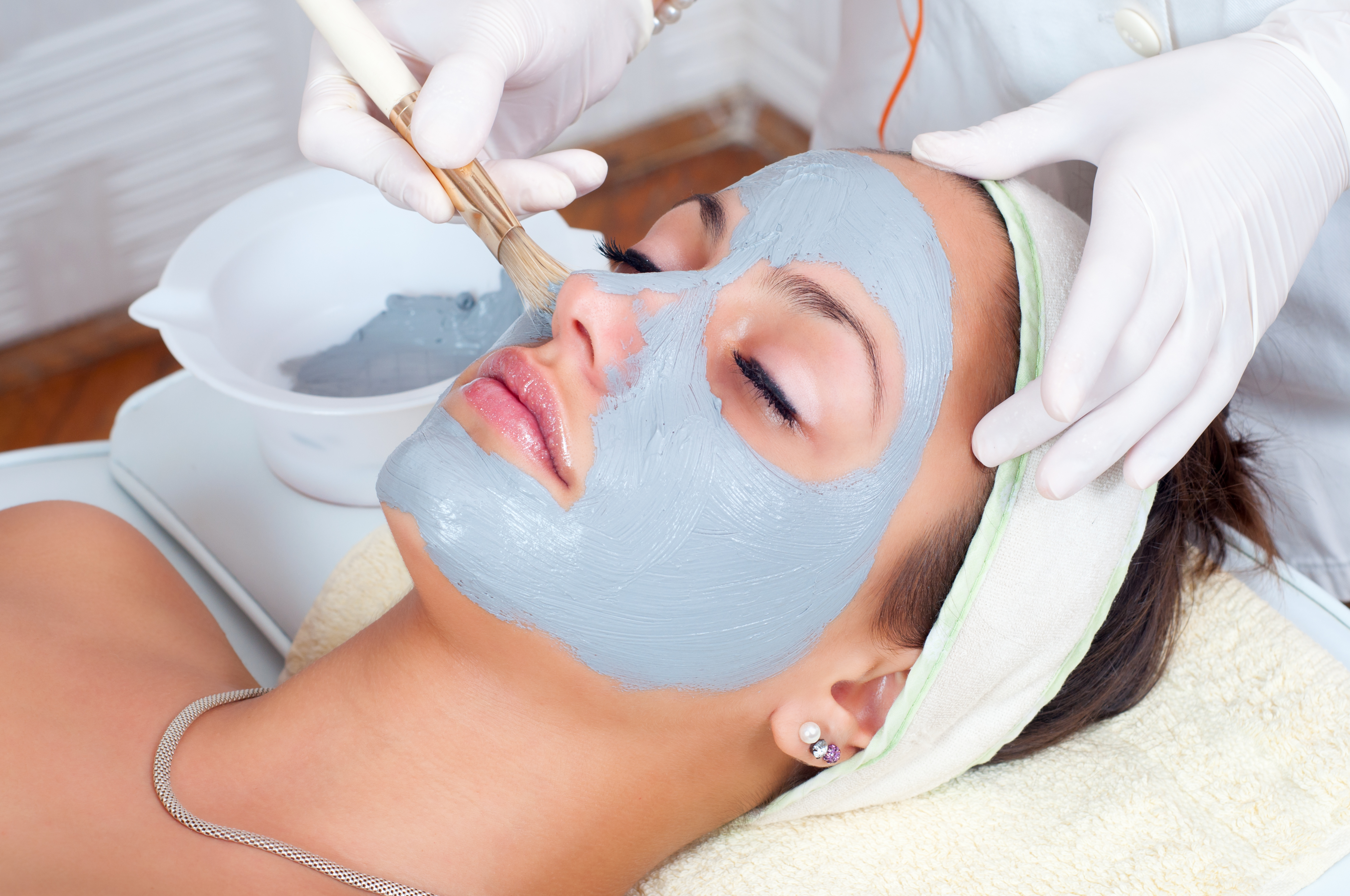 Young woman having blue facial mask applied by esthetician