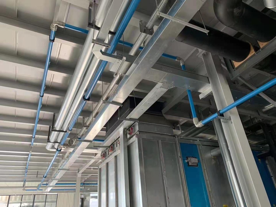 Complex network of metal airline pipes installed overhead in an industrial setting, featuring silver insulated and blue non-insulated pipes against a gray ceiling. The pipes are supported by a framework of steel beams and run alongside electrical conduits, suggesting a well-organized mechanical system within a manufacturing or processing facility.