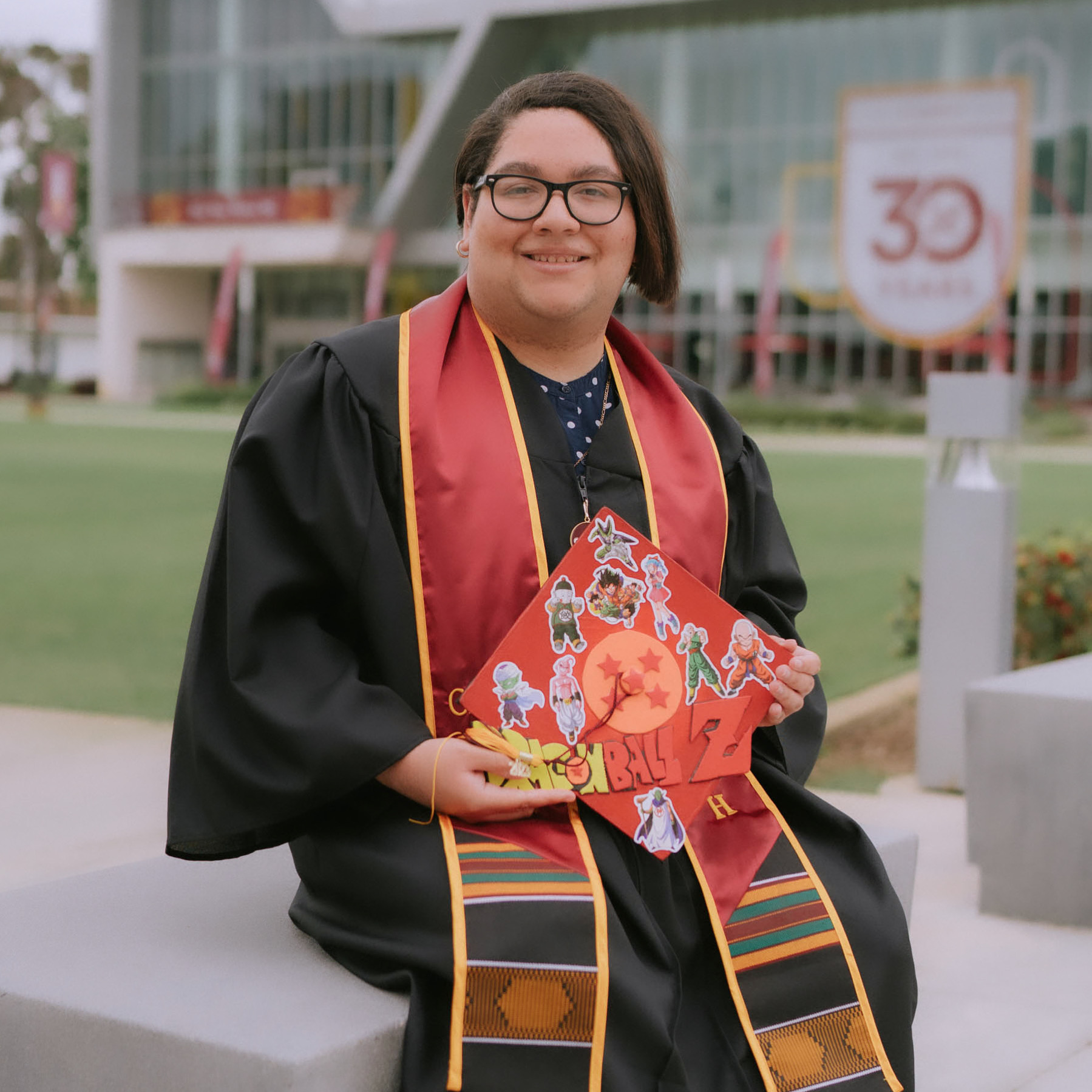 Biracial woman wearing a black graduation gown with a CSUDH sash. She is holding a Dragon Ball Z decorated cap while sitting down in front of the Loker Student Union.