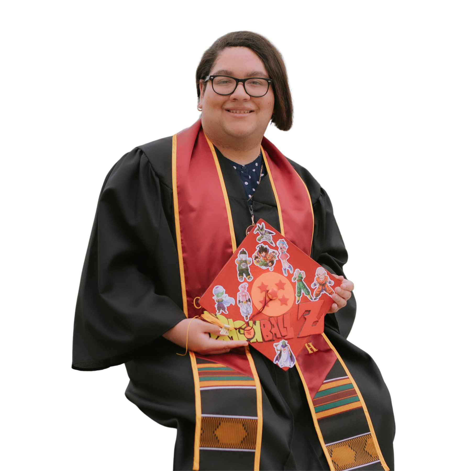 Black and Hispanic woman wearing a black graduation gown with a CSUDH sash. She is holding a Dragon Ball Z decorated cap while sitting down in front of the Loker Student Union.