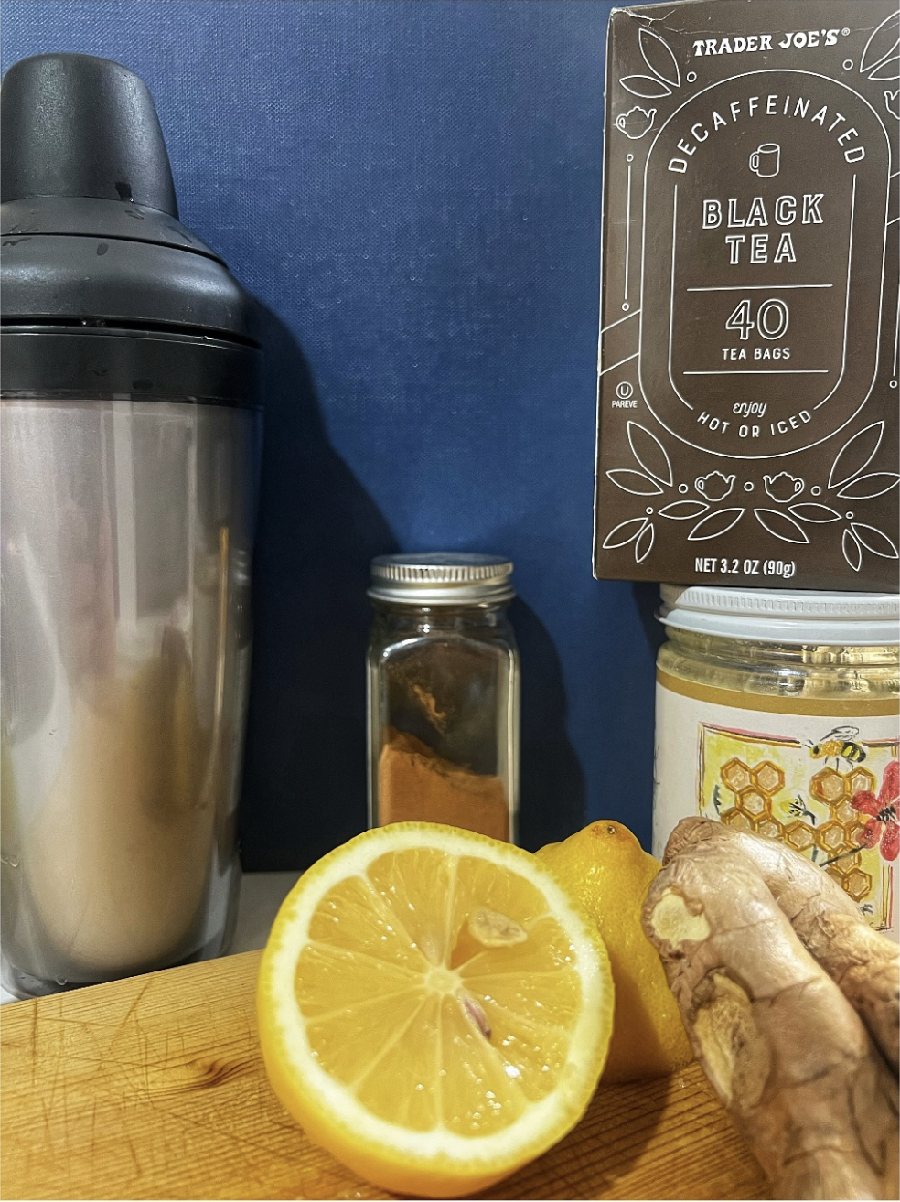 A close-up view of a kitchen setup featuring a shiny silver shaker, a box of Trader Joe's Decaffeinated Black Tea with 40 tea bags, a small jar of cinnamon, a glass jar of honey, and fresh ginger and lemon slices arranged on a wooden cutting board against a navy blue background.