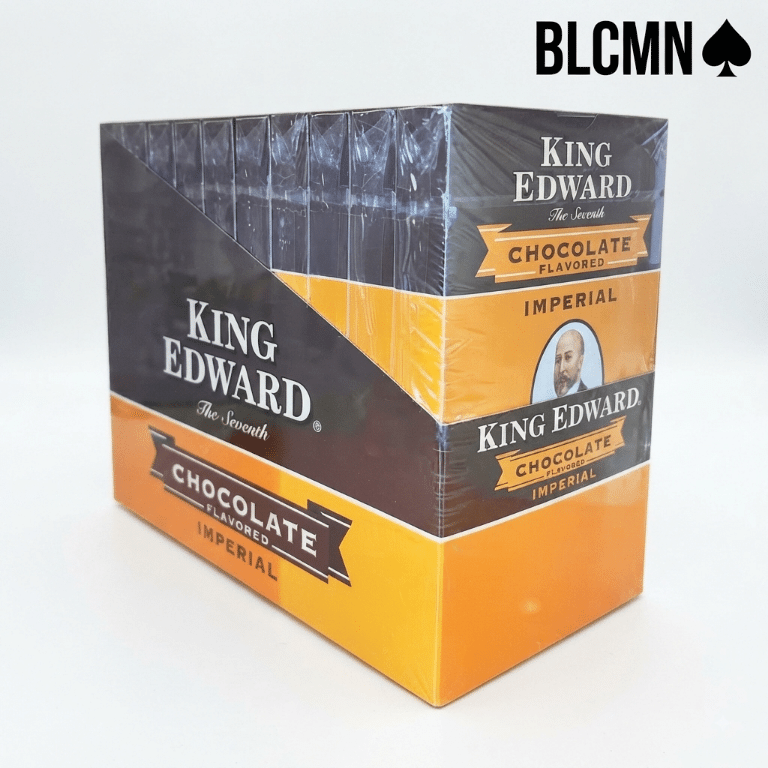 Studio product photograph on a clean white background showcasing an open master display box of King Edward Imperial Chocolate cigars filled with 10 individual retail packs, accompanied by a single cellophane-wrapped chocolate-flavored cigar standing upright beside the box. The black typography logo "BLCMN♠️" is placed in the top right corner.